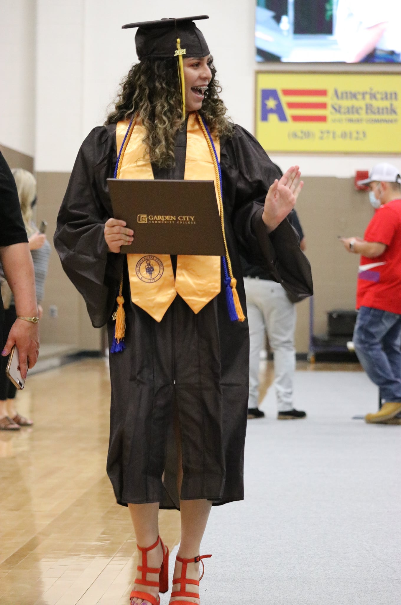 Graduate in cap and gown holding a Garden City Community College diploma and waving during a ceremony.
