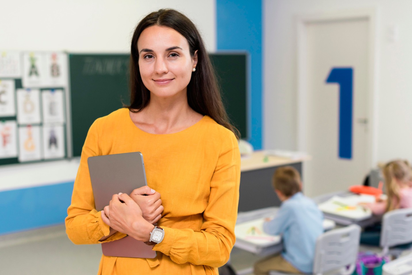 Person holding a tablet in a classroom while children work at desks in the background.