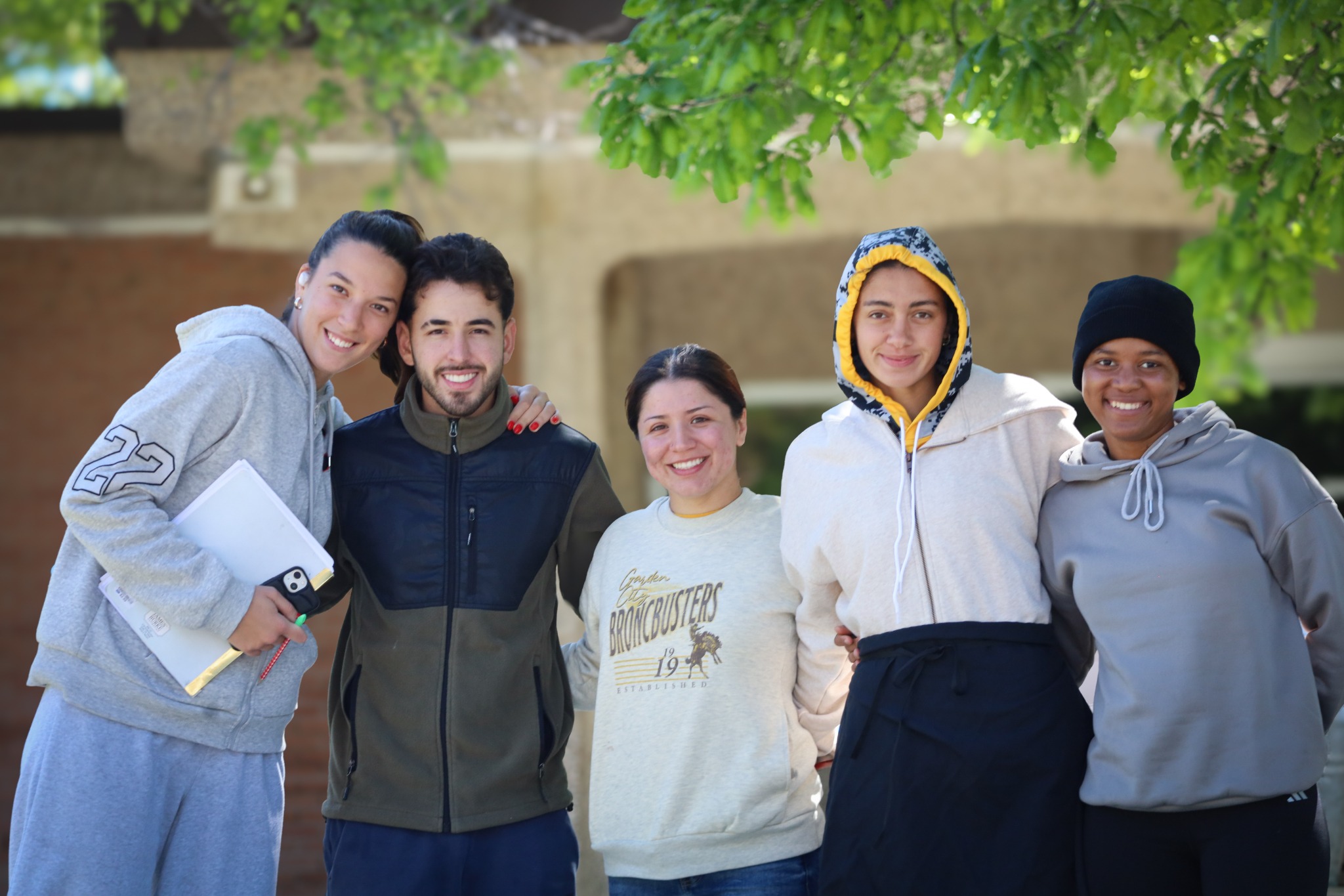 Group of students standing together outdoors on campus beneath a tree, posing with notebooks and backpacks.