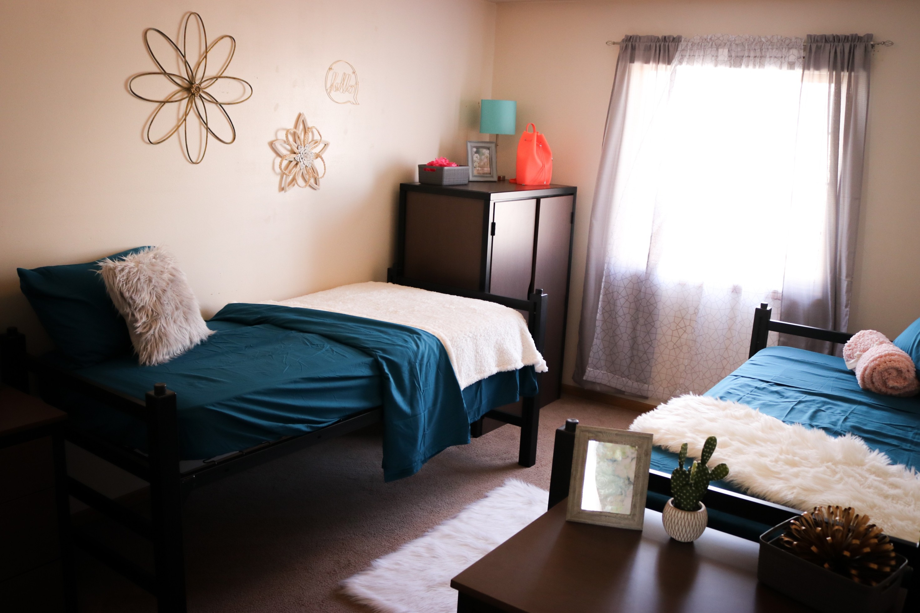 Bedroom with two single beds, teal bedding, dresser, and window with curtains letting in natural light.